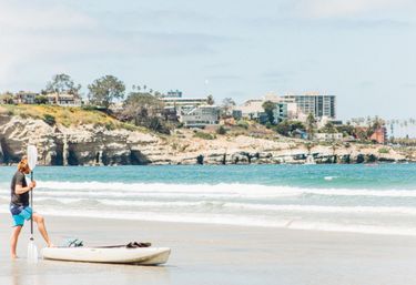 Person standing by a kayak holding a paddle on a sunny California beach, turquoise waves lapping the sand with rocky cliffs and a coastal town in the background.
