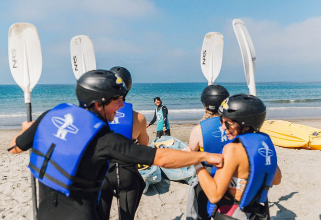 Excited group of kayakers on a sandy beach wearing blue life jackets and black helmets, holding white paddles while an instructor stands by colorful kayaks at the shoreline under a clear blue sky.