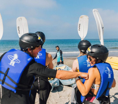 Excited group of kayakers on a sandy beach wearing blue life jackets and black helmets, holding white paddles while an instructor stands by colorful kayaks at the shoreline under a clear blue sky.