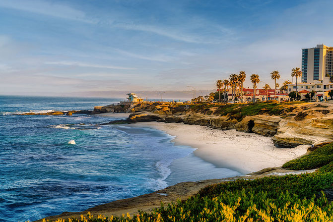 Sunlit Southern California coastline near San Diego with a sandy beach, rocky sandstone cliffs, palm trees, gentle waves and seaside buildings