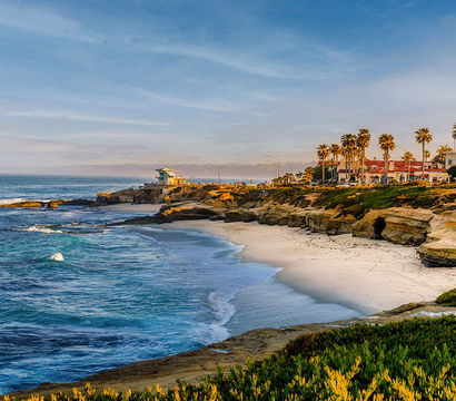 Sunlit Southern California coastline near San Diego with a sandy beach, rocky sandstone cliffs, palm trees, gentle waves and seaside buildings