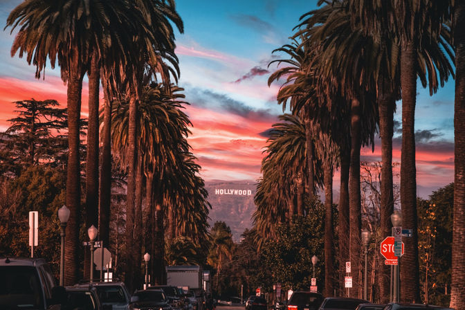 Palm-tree-lined Los Angeles street at sunset with a colorful sky and the Hollywood sign on distant hills.