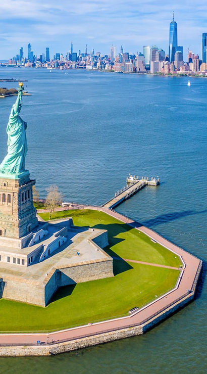 Aerial view of the Statue of Liberty on Liberty Island in New York Harbor with the Manhattan skyline across the water on a clear, sunny day.