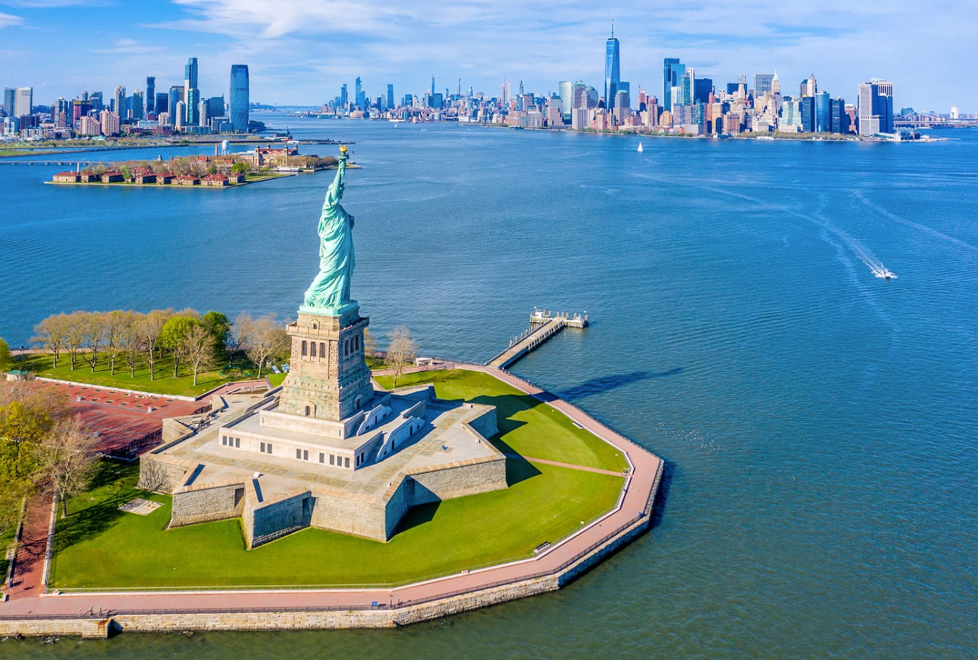 Aerial view of the Statue of Liberty on Liberty Island in New York Harbor with the Manhattan skyline across the water on a clear, sunny day.