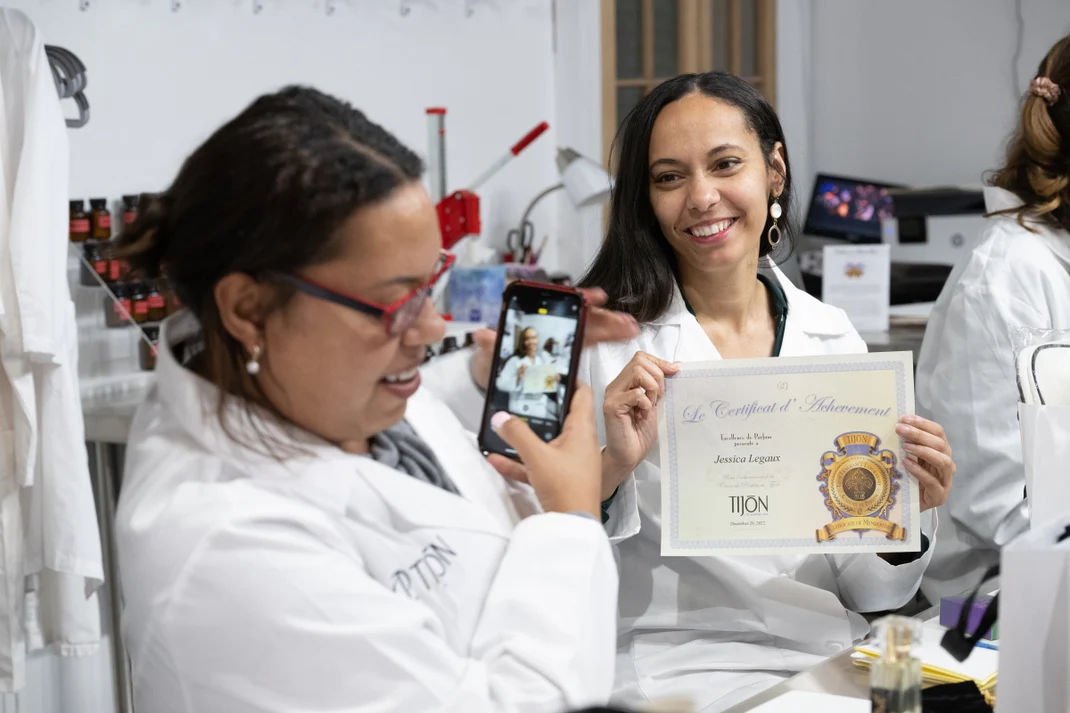 Smiling woman in a white lab coat holding a certificate while a colleague photographs her with a smartphone in a bright training lab or beauty workshop