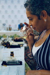 Woman sniffing a glass beaker at a fragrance tasting workshop, with amber sample bottles, scent strips and evaluation notes on the table.