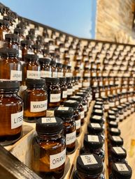 Curving scent-library wall of amber glass fragrance bottles with white labels (orange, mandarin, jasmine) neatly arranged on wooden shelves in an artisanal perfume studio
