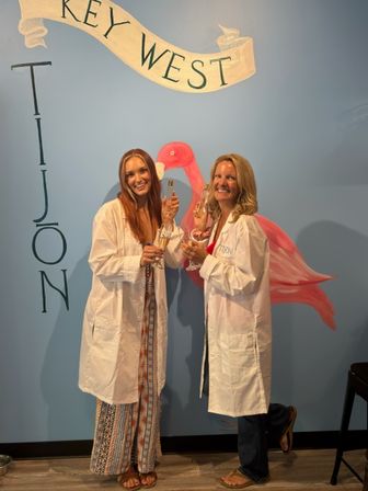 Two smiling people in white lab coats clinking champagne flutes in front of a blue Key West mural with a pink flamingo.