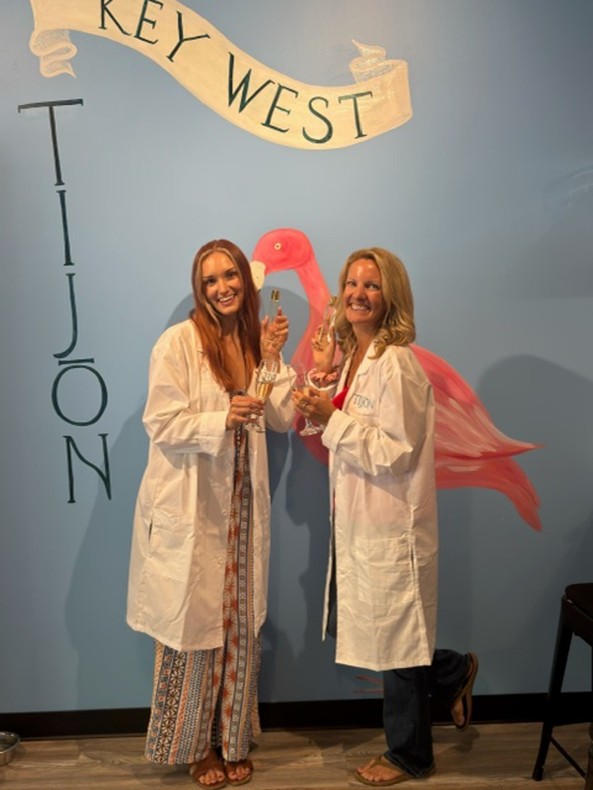 Two smiling people in white lab coats clinking champagne flutes in front of a blue Key West mural with a pink flamingo.