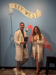 Two people in white lab coats pose in front of a blue Key West wall mural with a painted pink flamingo and banner, each holding small bottles — playful Key West photo.
