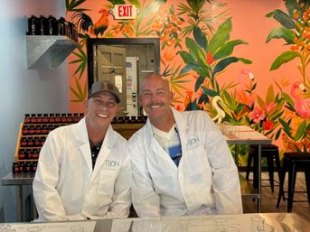 Two smiling people in white lab coats at a tasting counter with rows of small bottles, a vibrant tropical pink mural with plants and flamingos, and an EXIT sign overhead.