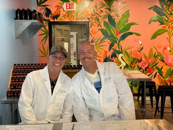 Two smiling people in white lab coats at a tasting counter with rows of small bottles, a vibrant tropical pink mural with plants and flamingos, and an EXIT sign overhead.