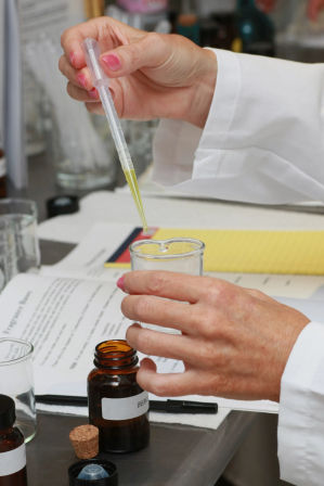 Close-up of hands in a white lab coat using a plastic pipette to drop yellow solution into a small glass beaker, with amber reagent bottles and notes on a laboratory bench