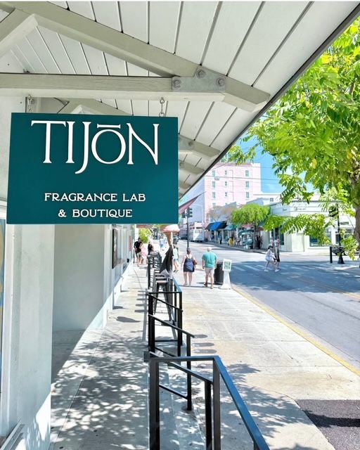 Teal hanging sign for a fragrance boutique under a white awning on a sunny, tree-lined main street — shaded sidewalk with railings and pedestrians passing storefronts.