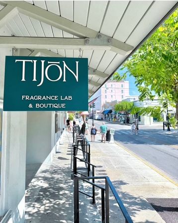 Teal hanging sign for a fragrance boutique under a white awning on a sunny, tree-lined main street — shaded sidewalk with railings and pedestrians passing storefronts.