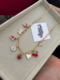 Playful gold link charm necklace in a beige jewelry box, with colorful enamel charms including a red sneaker, pacifier, white flower, round 'S' initial, strawberry, white heart, pair of cherries and a tiny animal face.
