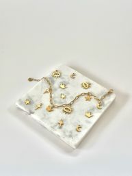 Gold charm chain necklace and scattered gold charms (sun, moon, stars, crowns) on a white marble slab — minimal jewelry flatlay on a clean white background.