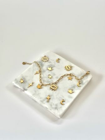 Gold charm chain necklace and scattered gold charms (sun, moon, stars, crowns) on a white marble slab — minimal jewelry flatlay on a clean white background.