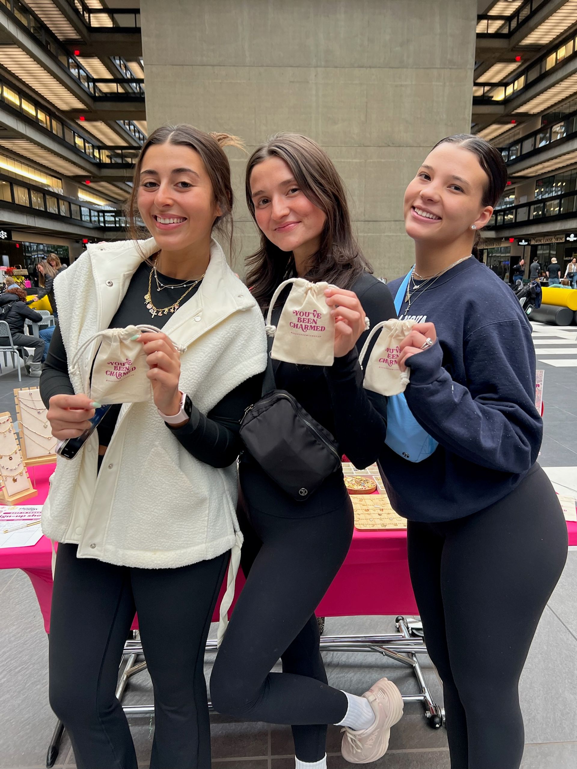 Three smiling women in a modern multi-level indoor atrium holding small drawstring pouches reading "YOU'VE BEEN CHARMED" at a jewelry pop-up table, dressed in casual athleisure and layered necklaces.