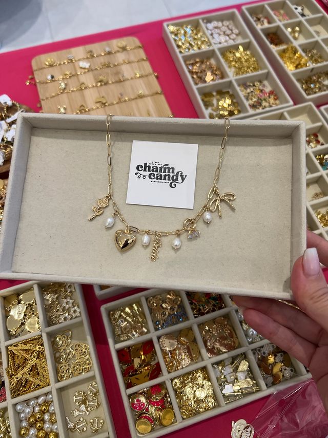 Tray held over a craft table showing a gold charm necklace with heart, pearls, rose and bow charms, surrounded by compartmented trays of gold charms and jewelry findings.