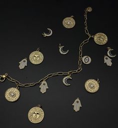 Flat lay of a gold charm bracelet and loose pendants—sun-face medallions, crystal-studded crescent moons, hamsa hands and an evil-eye disc—on a matte black background