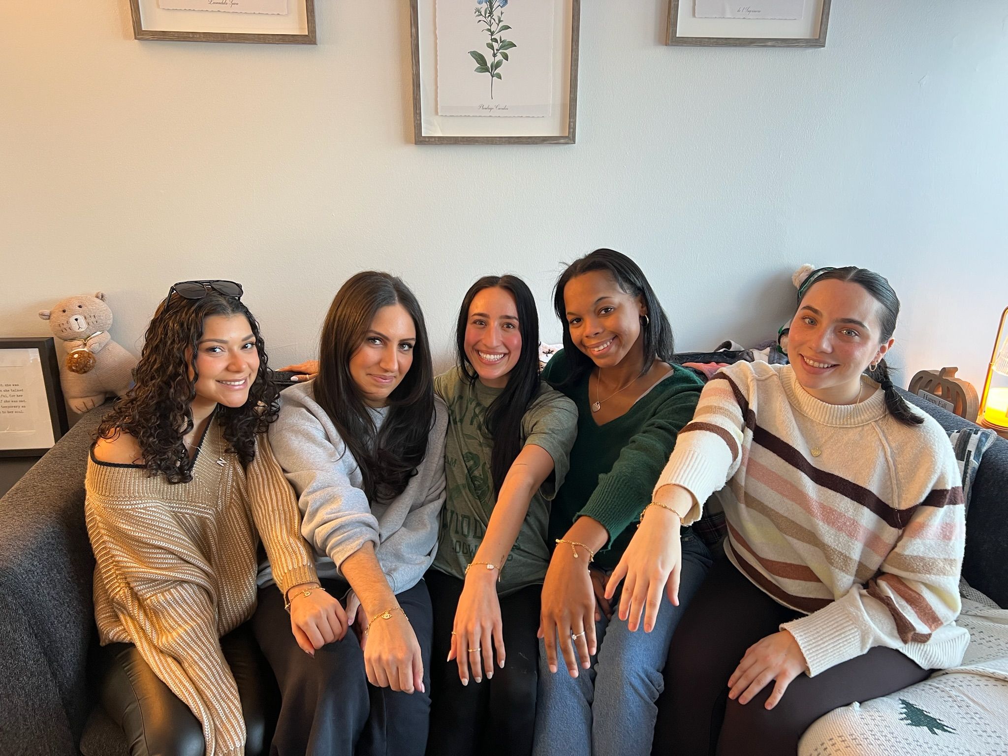 Five smiling friends seated on a gray couch in a cozy apartment living room, leaning forward to display rings and bracelets on their hands with framed botanical prints and a plush toy on the wall behind them.