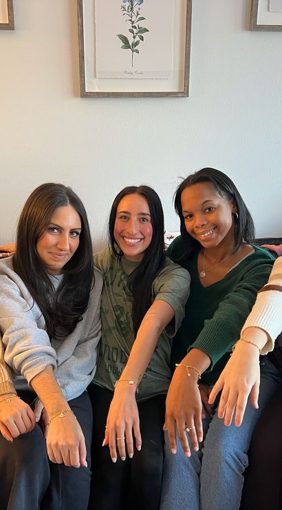 Five friends seated on a couch in a cozy living room, smiling and showing matching bracelets and rings, with framed botanical prints, a stuffed bear, and a warm lamp in the background.