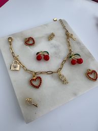 Flatlay of a gold charm bracelet and loose charms on a white marble slab — red enamel cherries, red-studded heart pendants, tiny gold lock and rhinestone key accents, styled jewelry photo