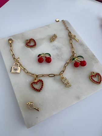 Flatlay of a gold charm bracelet and loose charms on a white marble slab — red enamel cherries, red-studded heart pendants, tiny gold lock and rhinestone key accents, styled jewelry photo