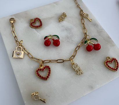 Gold charm bracelet with red rhinestone heart charms, enamel cherry charms, tiny padlock and key pendants arranged on a white marble tray