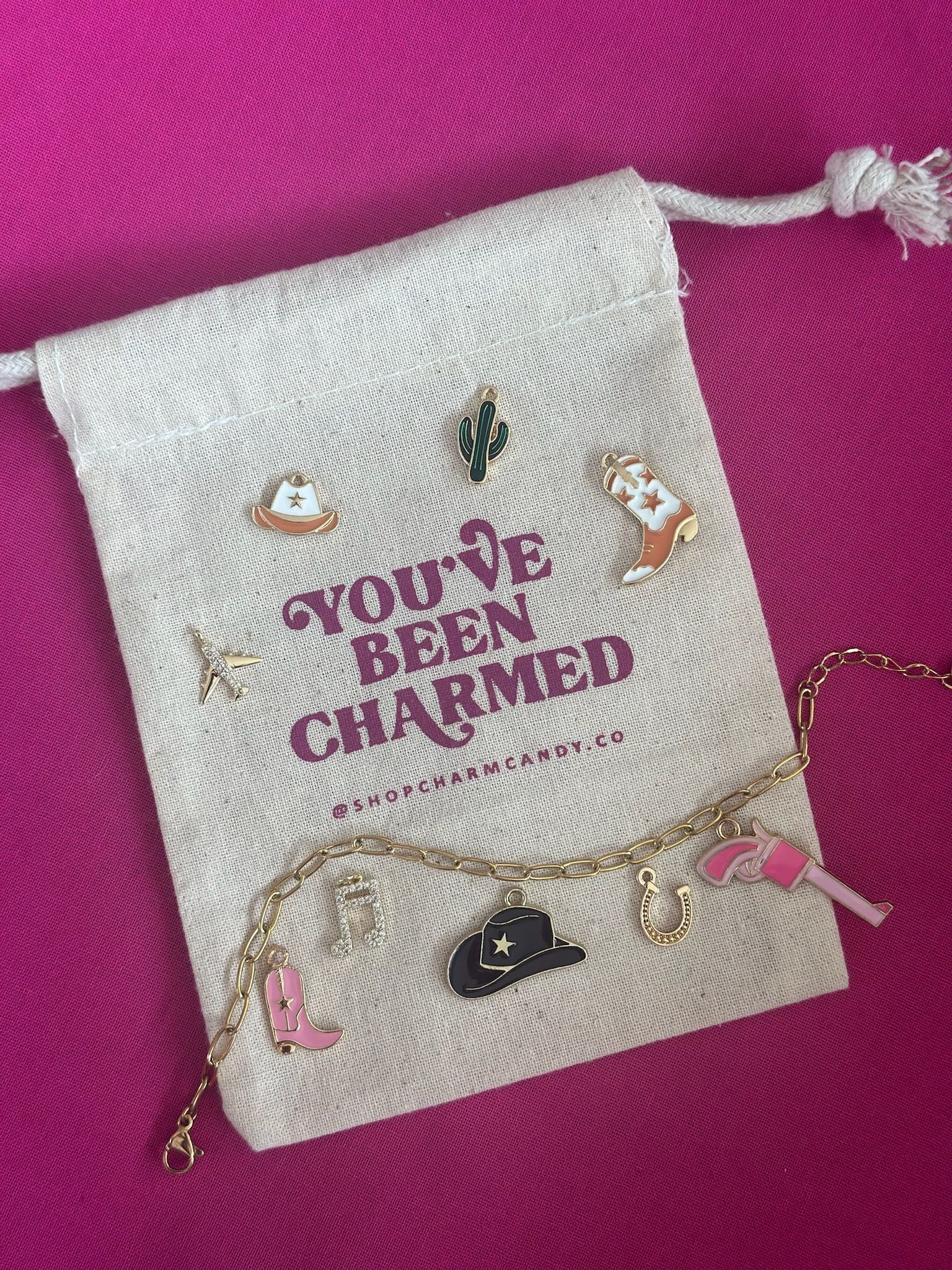 Cream drawstring pouch on bright pink background scattered with Western-themed enamel charms — cowboy hats (black and white), cowboy boots (pink and tan), a green cactus, gold horseshoe, small gold airplane, rhinestone music note and a gold chain bracelet.