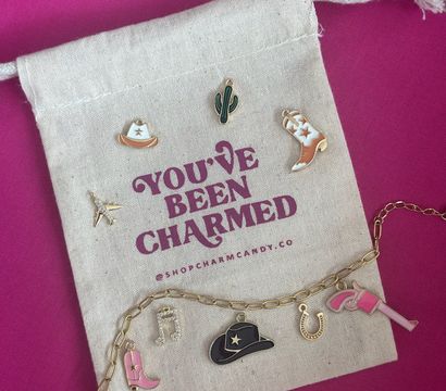 Enamel western-themed charms — cowboy hats, cowgirl boots, cactus, horseshoe and music note — arranged on a canvas drawstring pouch with magenta text and a gold charm chain on a bright pink background.