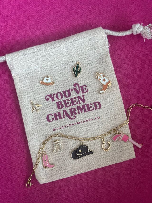 Enamel western-themed charms — cowboy hats, cowgirl boots, cactus, horseshoe and music note — arranged on a canvas drawstring pouch with magenta text and a gold charm chain on a bright pink background.