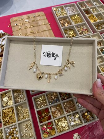 Gold charm necklace with heart, freshwater pearls and bow charms on a beige display tray held above divided trays of loose gold charms at a jewelry market booth