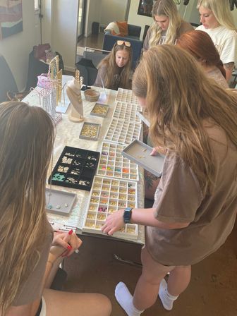 Group of women gathered around a living-room table at an at-home craft party, selecting beads, gold charms and necklace components from compartment trays and display stands to make DIY jewelry.