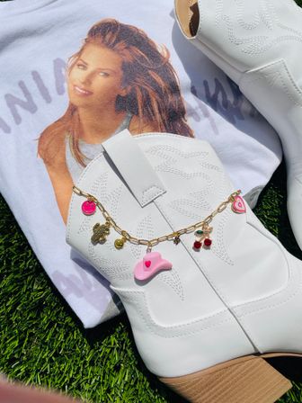 Flat-lay of white western cowboy boots with stitched detailing and a gold charm chain (pink cowgirl hat, cherries, smiley hearts) styled on a vintage photo graphic tee on green grass in bright sunlight