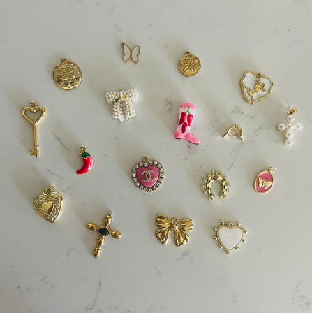 Flat-lay of assorted small gold and enamel jewelry charms—hearts, key, bow, horseshoe, pearl cross, red chili and pink cowboy boot—playful arrangement on a light marble countertop.