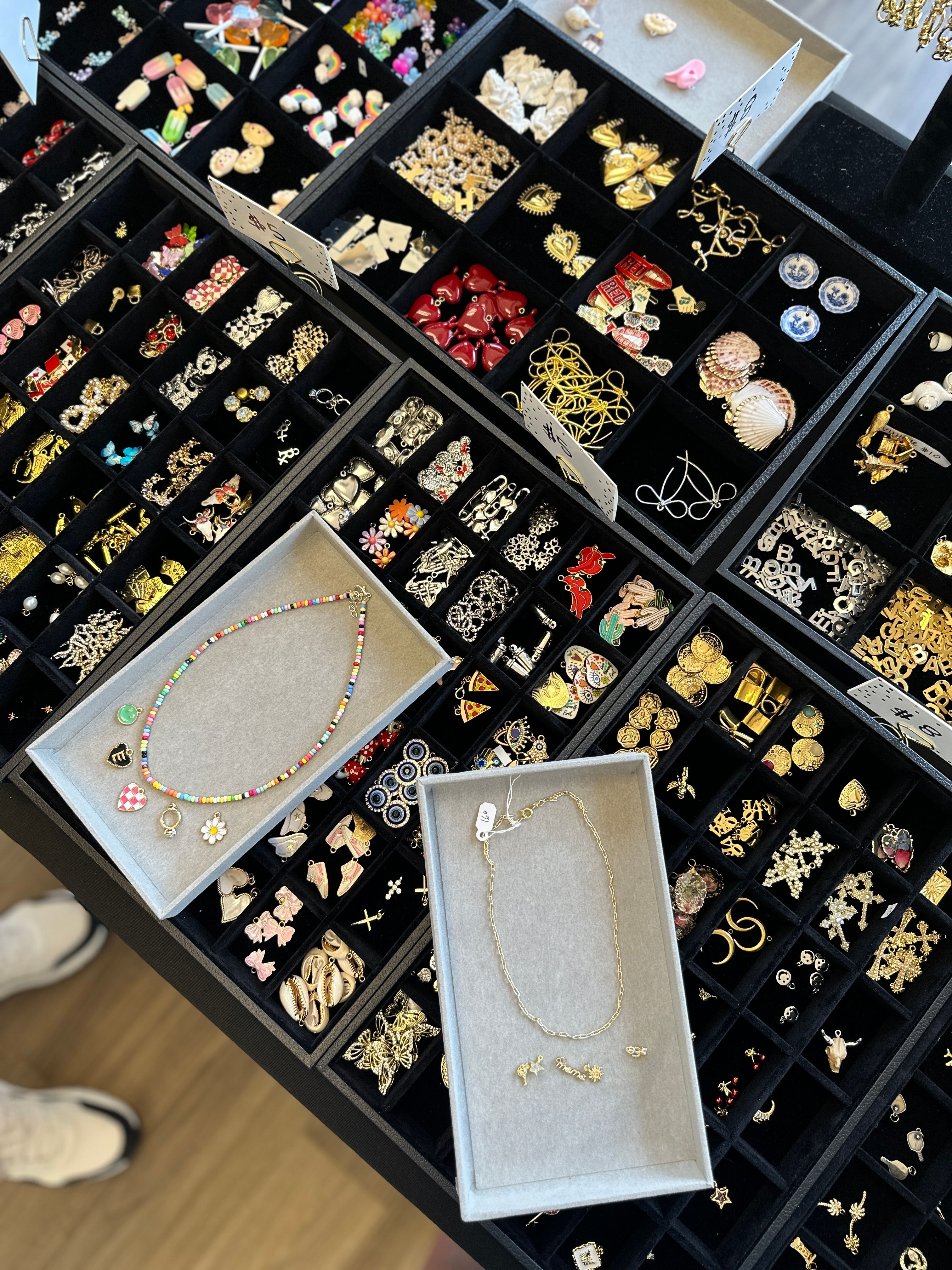 Top-down view of a tabletop jewelry display with black compartment trays filled with assorted gold and silver earrings, charms, pins and novelty pieces; two gray trays showcase a colorful beaded necklace with charms and a delicate gold chain.