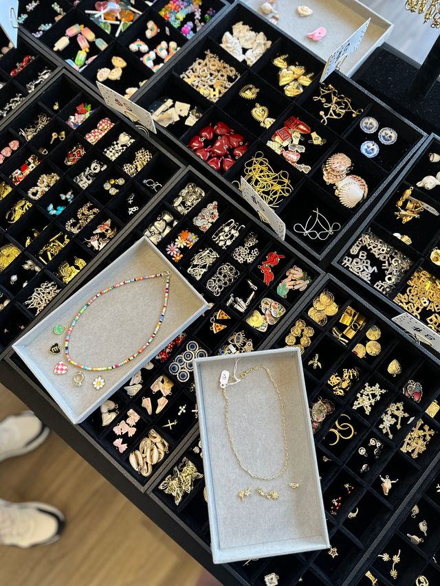 Top-down view of a tabletop jewelry display with black compartment trays filled with assorted gold and silver earrings, charms, pins and novelty pieces; two gray trays showcase a colorful beaded necklace with charms and a delicate gold chain.