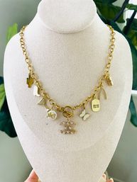 Gold charm chain necklace displayed on a beige velvet bust — mixed charms including cactus, boot, martini glass, pearl 'Mom' bead, sparkling rhinestone double-loop pendant, white enamel butterfly, '11:11' tag, wishbone and mini coffee cup, photographed with green plant background; women's fashion jewelry and gift idea.