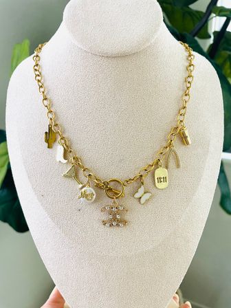 Playful gold charm necklace on a beige display bust featuring assorted charms — cactus, cowboy boot, martini glass, pearl 'Mom' charm, sparkling logo pendant, butterfly, 11:11 tag and coffee cup.