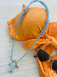 Summer flat-lay: textured orange bikini top tied in a knot, blue braided cord necklace with a pale fish pendant, tiny shell and pearl charms, and gold-rimmed sunglasses on a light wood surface.