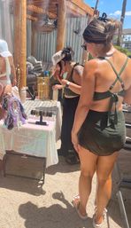 Poolside cabana pop-up with women in swimsuits browsing a table of gold necklaces, bracelets and colorful hair scarves under a wooden pergola on a sunny resort day.