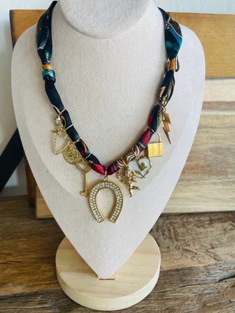 Boho-chic statement scarf necklace on beige velvet display bust and round wooden stand, twisted colorful silk ribbon with gold charms — rhinestone horseshoe pendant, key, lock, lightning bolt, cherub and flower — women's fashion jewelry on rustic wood table