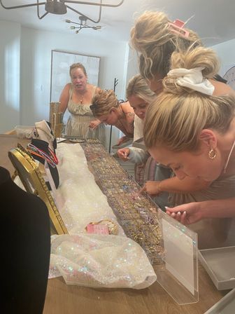 Several women leaning over a sparkly sequined table inspecting trays and clear organizers of rings, necklaces and colorful jewelry at a casual home jewelry party.