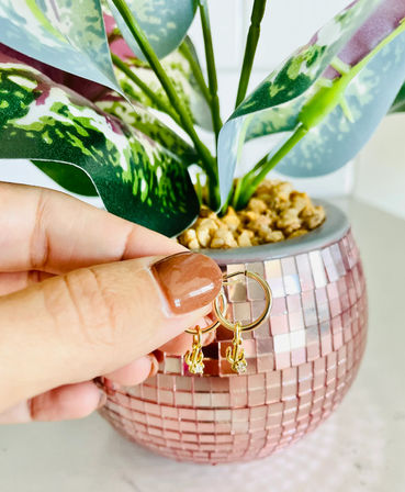 Hand holding gold hoop earrings with tiny cactus charms in front of a pink mosaic planter and variegated houseplant on a white tabletop