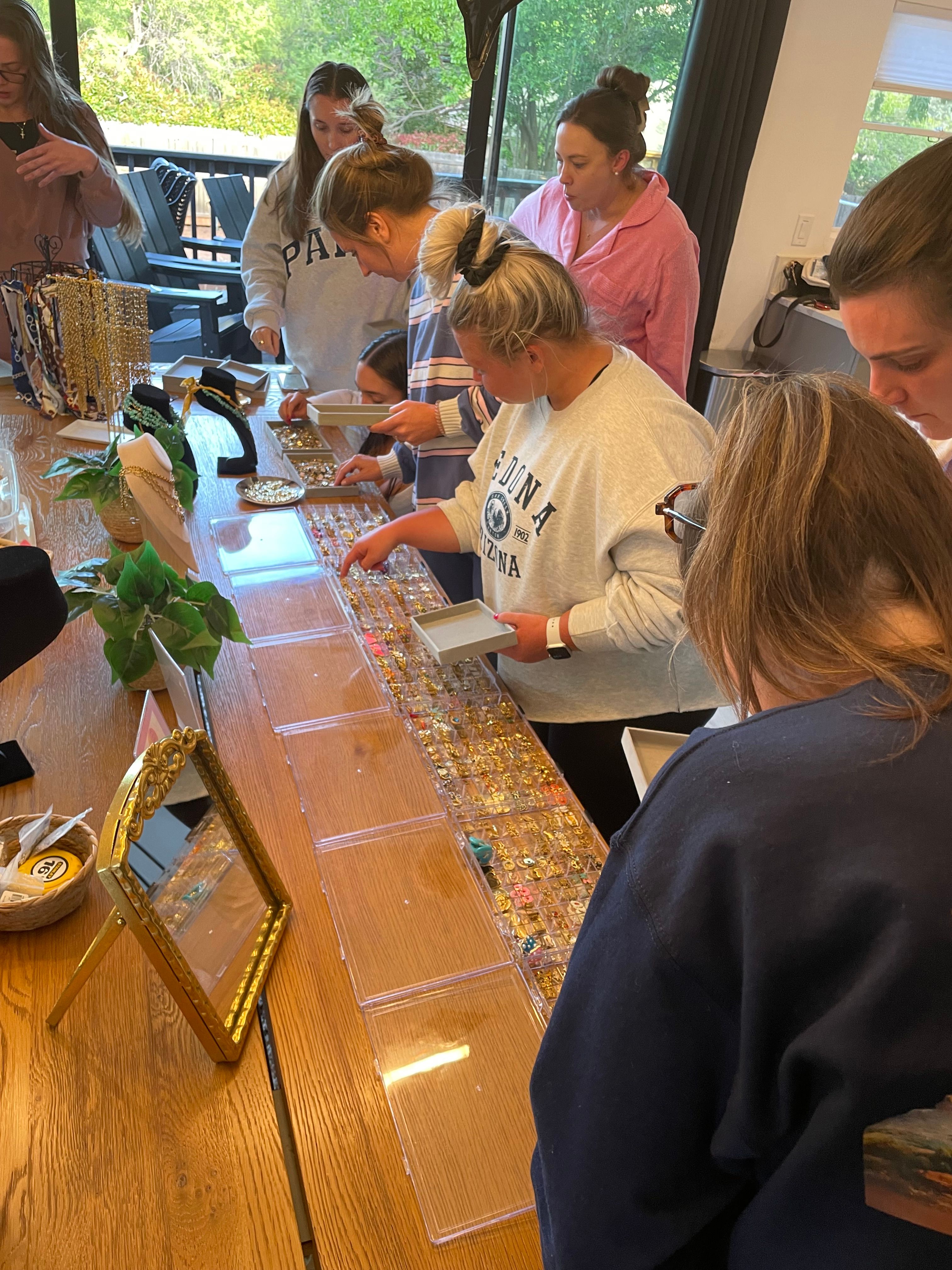 Group of women browsing a tabletop jewelry display—clear trays of rings, earrings and necklaces on a wooden table at an indoor shopping party.