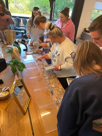 Group of women browsing a tabletop jewelry display—clear trays of rings, earrings and necklaces on a wooden table at an indoor shopping party.