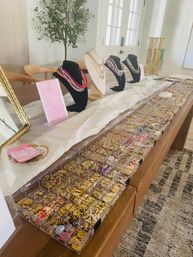 Bright indoor jewelry display on a wooden table in a home entryway: busts showcasing layered necklaces, racks of gold chains and clear organizers filled with charms, earrings and beads.