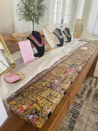 Bright indoor jewelry display on a wooden table in a home entryway: busts showcasing layered necklaces, racks of gold chains and clear organizers filled with charms, earrings and beads.